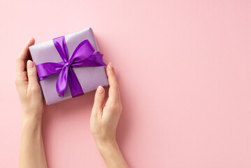 New Year concept. First person top view photo of woman's hands holding violet giftbox with purple ribbon bow on isolated pastel pink background with copyspace