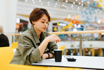Pretty asian woman with short hair drinking hot drink while sitting at a table in the mall food court cafe