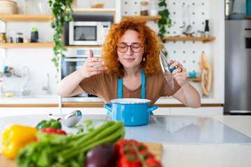Cheerful young pretty female in apron prepare lunch and smell dish in kitchen at home. Woman cooking dinner for family at new recipe at home.