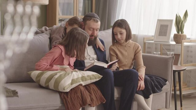 Jewish man reading the Hebrew Bible to his little daughters on Hanukkah, sitting together on sofa in living room