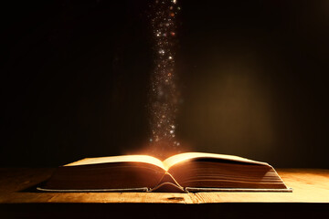 Magical image of open antique book over wooden table with glitter overlay