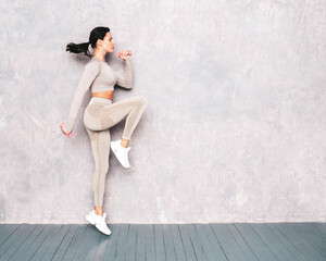 Fitness woman in sexy sports clothing. Young beautiful model with perfect body. Female posing in studio near grey wall at summer sunny day. Jumping and running. Doing exercises with dumbbells