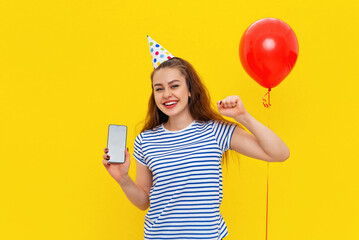 Wow, I win. Portrait of amazed woman holding cell phone with empty screen for ad, wears party cone on head shouting in surprise, shocked by winning, standing over yellow background