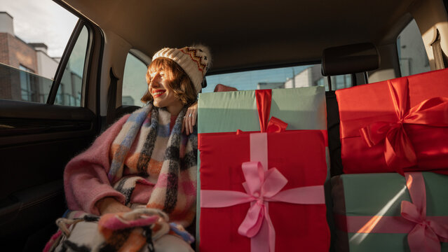 Young woman dressed for the holidays drives in car on back seat with beautifully wrapped Christmas presents. In anticipation of the winter holidays and shopping. Wide angle shot