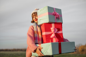 Young woman dressed for the holiday carries New Year's gifts outdoors on nature during sunset. In...