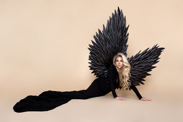 Stunning blonde woman in elegant long black dress and big wings is posing in studio. Luxury evening...