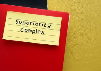 Red notebook on yellow background with handwritten text SUPERIORITY COMPLEX, means belief that your...