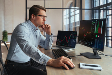 Side view of crypto broker sitting at office table, analyzing market, looking at screen of personal computer with candlestick chart, touching chin with hand, thinking, wearing shirt and glasses