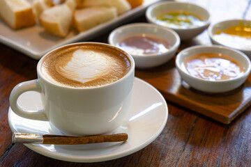 Latte art coffee in white cup on wooden desk with dessert and bread in the background