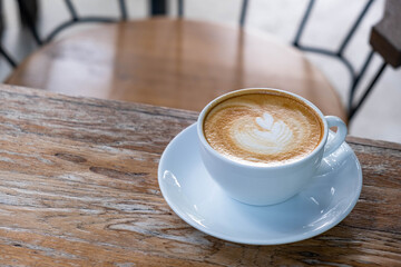 Latte art coffee in white cup on wooden desk with metal chair in the background with copy space