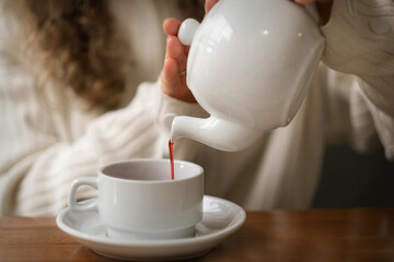 White blonde girl in a warm sweater drinks tea in a coffee shop. In a cafe, coffee is poured into a white mug. Hot tea for morning breakfast. Warming coffee in the restaurant on a winter evening. High