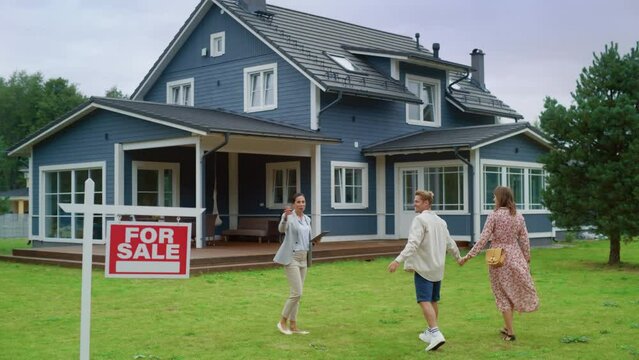 Establishing Shot: Professional Realtor Showing a For Sale Property to a Young Successful Couple. Female Real Estate Agent with Tablet Walking with Young Family to the House. Homeowners Concept.