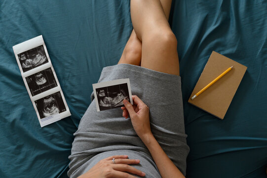 High angle shot of a pregnant woman lie down looking at a sonogram or ultrasonography picture while touching her belly in bedroom