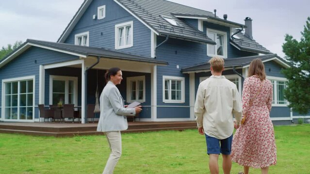 Real Estate Agent Showing a Beautiful House to a Young Happy Couple. People Walking Outside on a Lawn, Talking with Businesswoman, Discussing Buying a New Home. For Sale Sign on the Street.