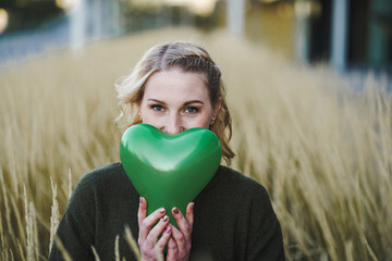 thoughtful young vegan woman holding a green heart air balloon in her hand infront of her face to...