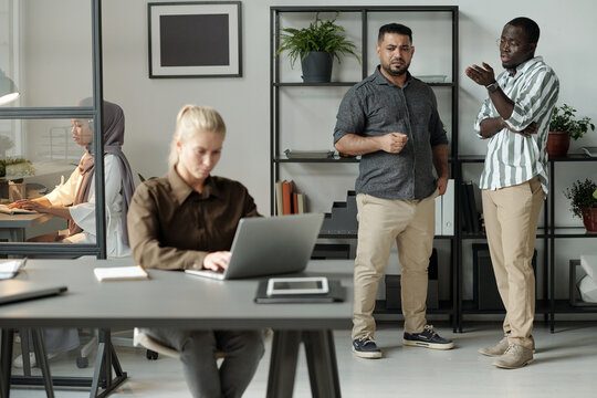Two intercultural men in casualwear bullying blond Caucasian woman with laptop sitting by desk in front of them and networking