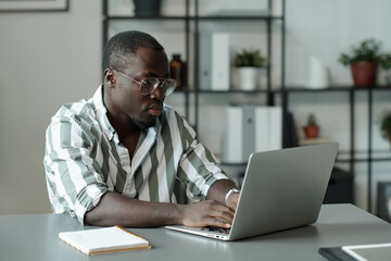 Young serious man in eyeglasses and striped shirt looking at laptop screen while sitting by workplace and preparing presentation