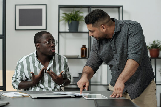 Middle aged boss bending over workplace of African American male subordinate while rebuking him for not carrying out working task