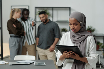 Young Muslim businesswoman in hijab holding black folder with documents against group of intercultural colleagues mocking at her - obrazy, fototapety, plakaty