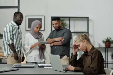 Obraz premium Young blond businesswoman touching head while sitting by workplace in front of laptop against group of intercultural colleagues