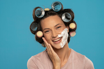 attractive happy  woman with hair curlers rollers posing with shaving foam on her face on a blue...