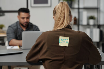 Back of young blond businesswoman with word loser written on sticky notepaper sitting by workplace...