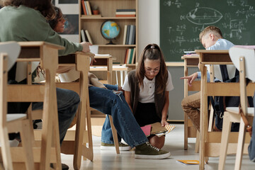 Cruel schoolkids laughing at their classmate standing on knees in aisle between desks and picking...