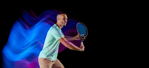 Movements in neon glow. Active male tennis player in sportwear playing tennis isolated over dark background in mixed purple neon light.