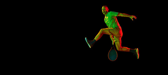 Studio shot of professional tennis player jumping with tennis racket isolated over dark background in neon light. Concept of professional sport, fashion, ad