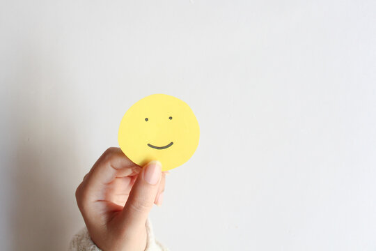 Woman hands holding yellow smile face paper on white background, top view, good feedback rating,think positive, customer review, assessment, world mental health day, Compliment Day.