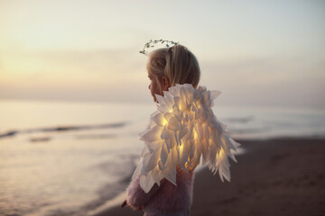 Cute little child girl standing on the sand beach near the sea during sunset with led glowing wings - 543182988