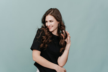 Attractive shy brunette girl toothy smiling looking aside twirls her curly long hair. Hispanic model posing at studio over turquoise backdrop. Playful young woman dancing indoors. Beauty and fashion.