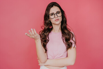Brunette young woman in glasses, pink t-shirt looks at camera with asking face expression shaking hand, asks. Attractive hispanic girl hesitating, makes choice stands over pink studio backdrop. Mockup