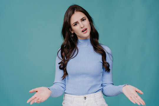 Disappointed brunette young woman with long wavy hair dressed in lilac sweater spreads hands in perplexity, upset stands over turquoise studio background. Peoples honest emotions. Mockup