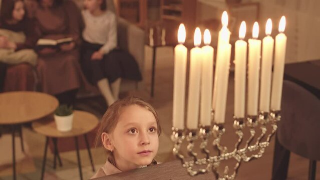 Little girl looking with interest at lit menorah candles standing in dark living room at Hanukkah night