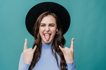 Playful brunette female hipster in hat with brim and lilac sweater shows tongue and rock n roll...