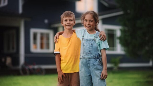 Young Cheerful Siblings Posing Outdoors, Hugging and Looking at Camera and Smile. Two Boy and Girl Friends Standing on a Lawn in Front of Suburban House, Enjoying Summer Time, Childhood, Friendship.