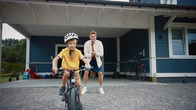 Cheerful Father Teaching His Son to Ride a Bicycle in The Front Yard of Their Beautiful Residential Area Home. Boy Wearing a Helmet, Bravely Balancing Himself on a Bike. Handheld Footage.