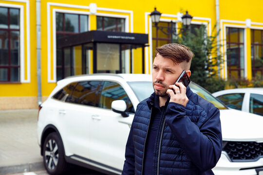 Frowning Tense Sullen Or Sad Concentrated Young Caucasian 34 Years Old Man With Goatee Beard Talks On Smartphone On Street Near White Car. Waist Up Lifestyle Business Portrait, Difficult Conversation