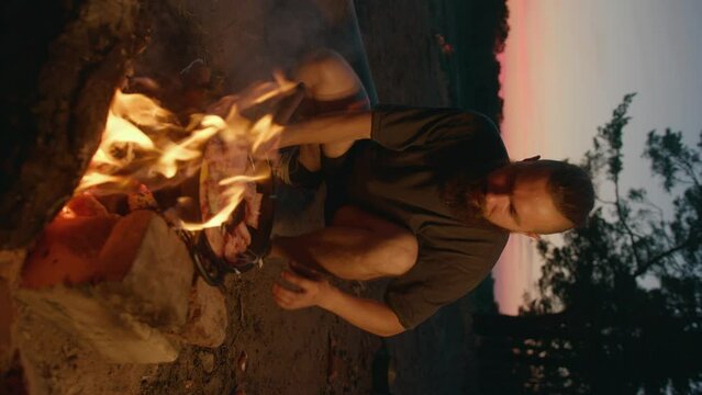 Young hipster man cooks food on fire on the river bank and breaks eggs into afrying pan with meat. Slow motion. Vertical video