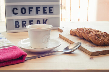 Close up view of a coffee cup, spoon, kitchen placemat, brown sugar, biscuits and letter board on a wooden table