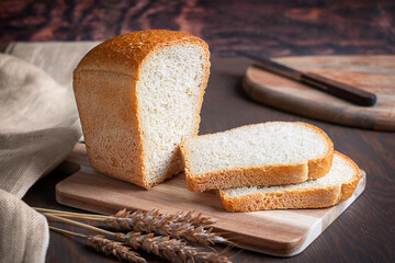 Homemade natural bread cooked from dough of wheat flour and yeast by baking, sliced and served on cutting board with ripe dry grains and towel on dark brown wooden table at kitchen for breakfast