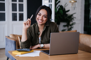 Cheerful young African American young woman sitting at desk with laptop and calculator, holding pen, looking at camera, toothy smiling. Satisfied pretty Brazilian entrepreneur satisfied by work.