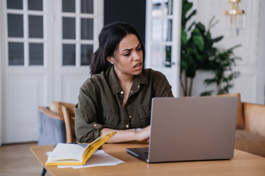Dissapointed young African American woman in shirt sitting at table with laptop makes video call, upset by  financial troubles. Tired businesswoman upset talking via internet. Business and finance.