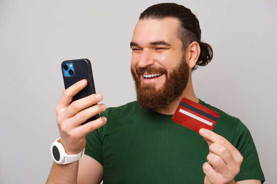 Image of happy handsome man with beard buying something online with credit card and smartphone.