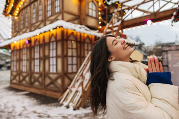 Smiling young caucasian girl laughing with closed eyes walks at amusement park during New Year. Brunette wavy hair woman wears sweater and jacket. Positive emotins, fun concept.