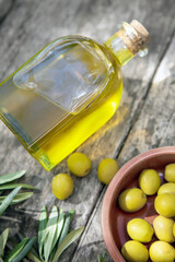 A glass bottle of olive oil lit by the sun, sprigs of olive and a plate of olives are on a wooden table. A vertical image.