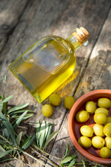 A glass bottle of olive oil lit by the sun, sprigs of olive and a plate of olives are on a wooden table. A vertical image.