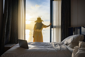 Laptop on white bed sheets in cozy room. Young businesswoman standing at window in confident pose...