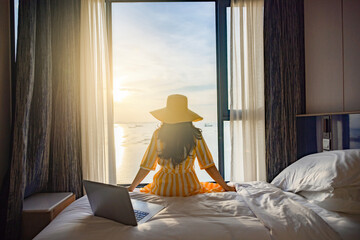Tourist woman sitting on bed nearly window, looking to Beautiful sunset above sea or ocean,...